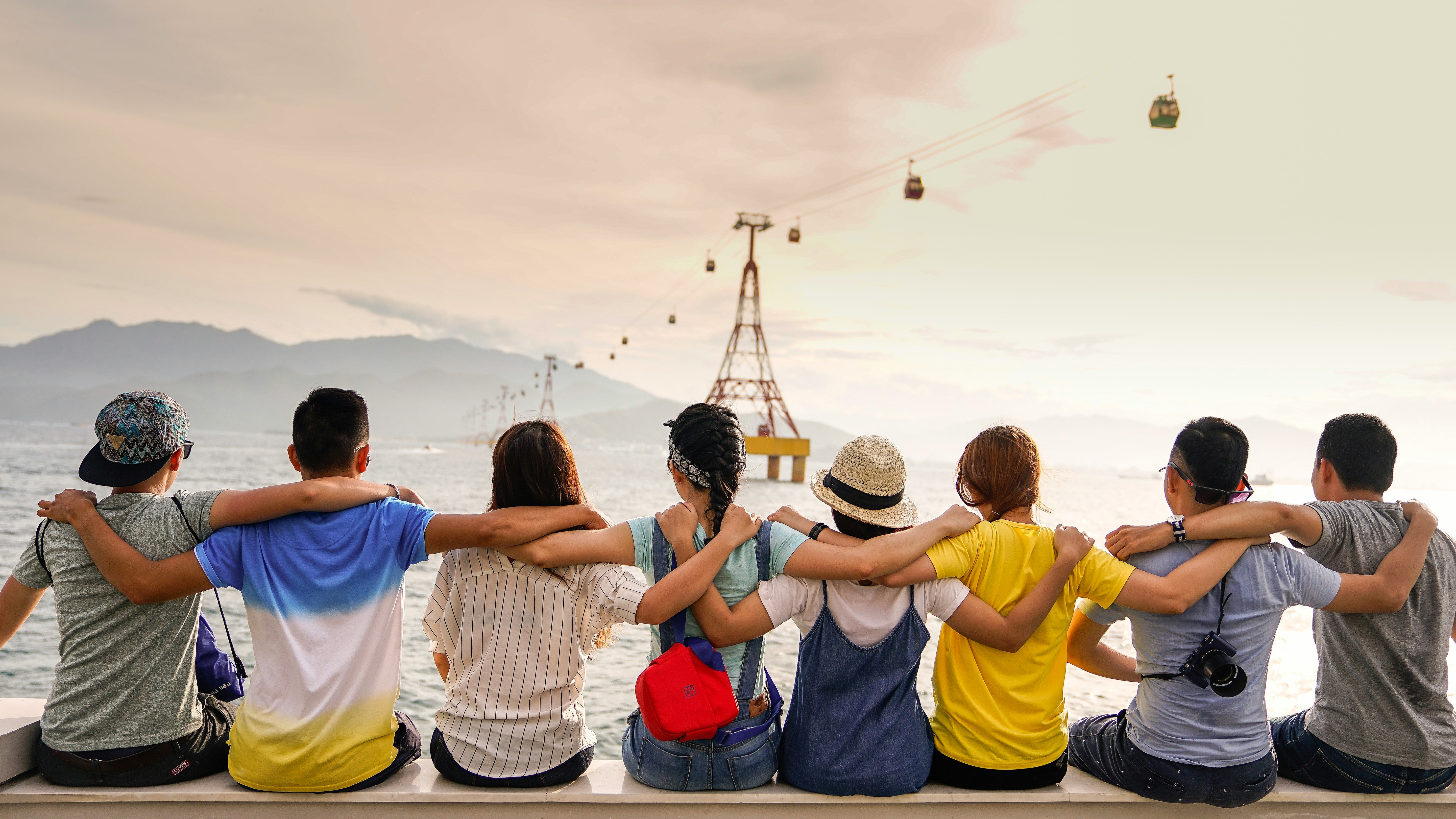People dressed in colorful attire with arms slung over each other's 
			shoulders sit facing away from the camera looking at an aerial tramway. 
			The picture has mountains in the distance.